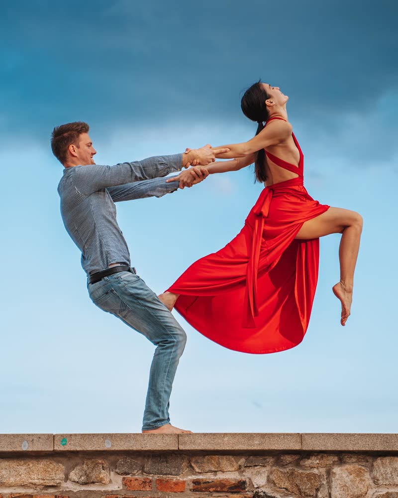 Dancer in a red dress leaning back in a counterbalance with her partner on a stone wall under a blue sky