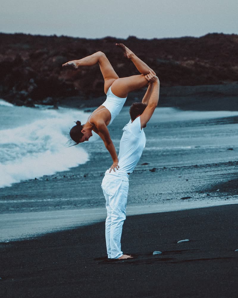 Acrobatic back-bend lift in white outfits on a black sand beach with waves