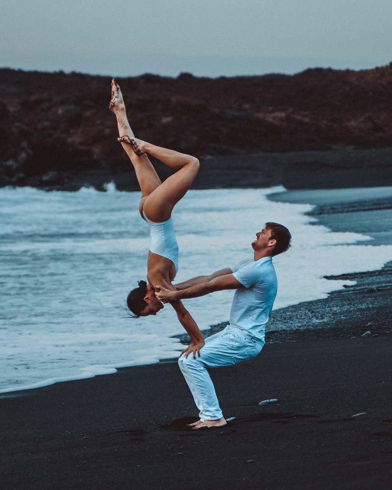 Inverted split balance supported by the partner in a deep squat on a black sand beach