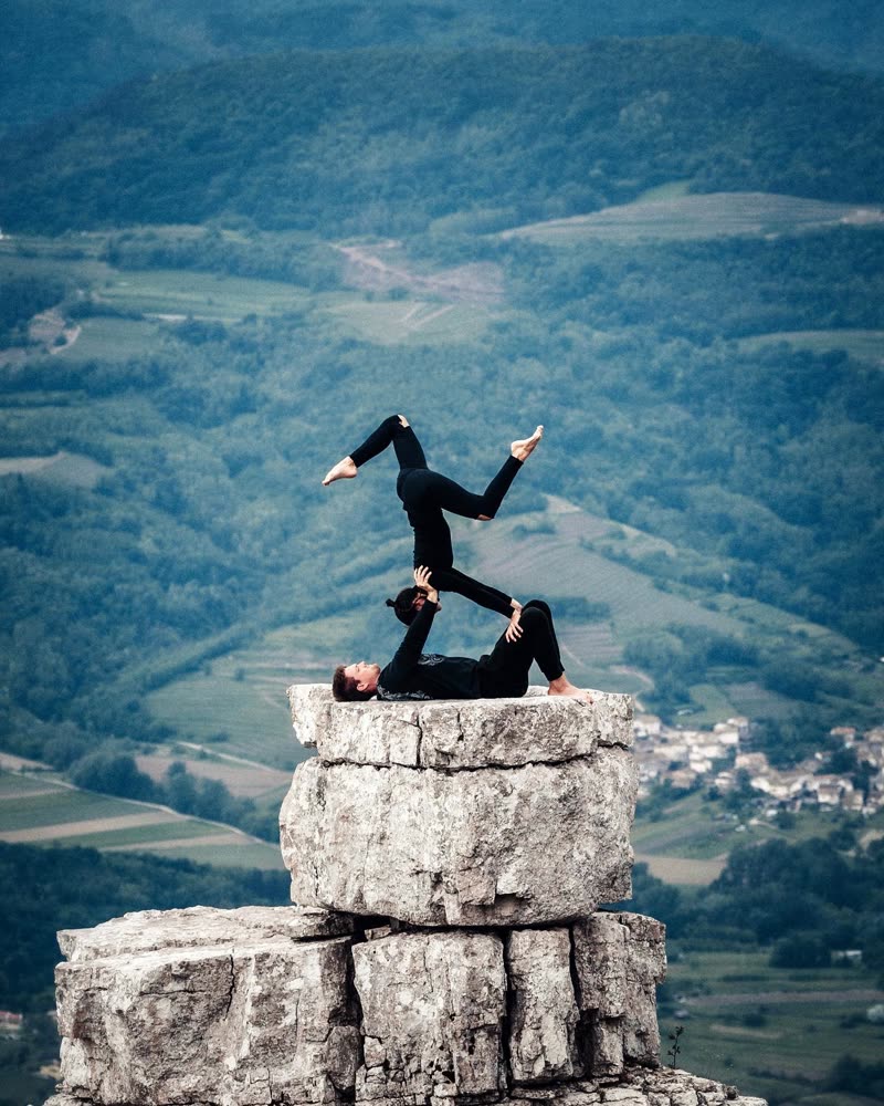 Two acrobats balancing in an inverted figure on top of a rock formation above a valley