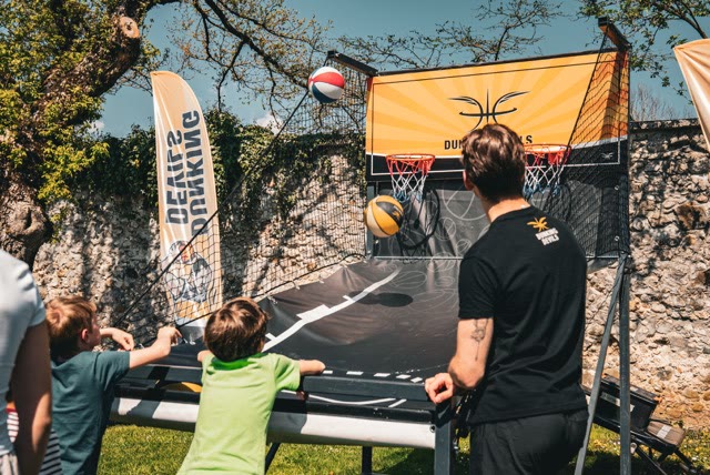 Children shooting at the arcade basketball game in a garden next to a Dunking Devils coach