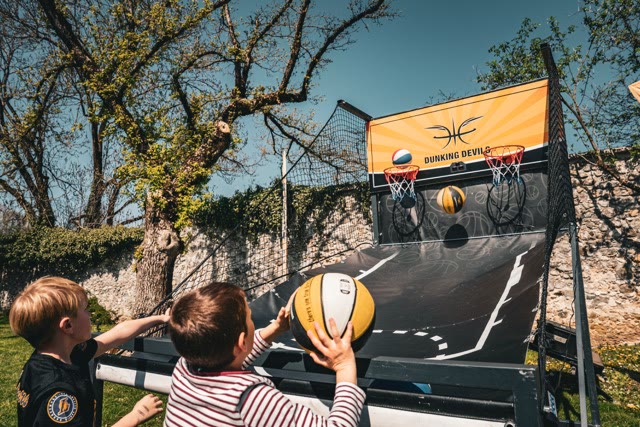 Two children playing arcade basketball, one holding a large yellow ball