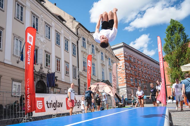 Young athlete doing a flip above a blue air track on a town square with spectators around