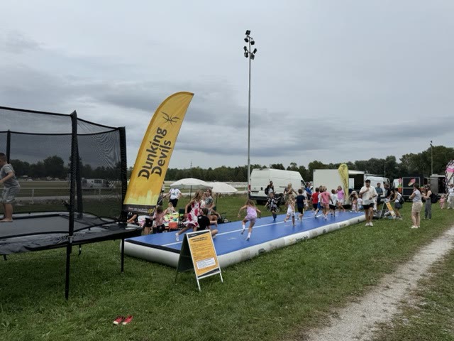 Children running along a long blue air track at an outdoor event with a Dunking Devils flag