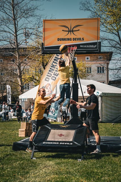 Child jumping from a mini trampoline to dunk on a Dunking Devils hoop at an outdoor fan zone, with two coaches spotting