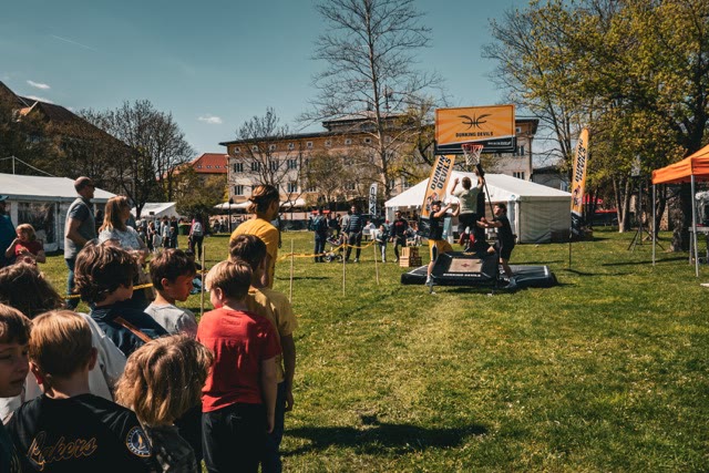 Children queuing for the basketball dunk workshop on a sunny park lawn with the hoop and trampoline in the background
