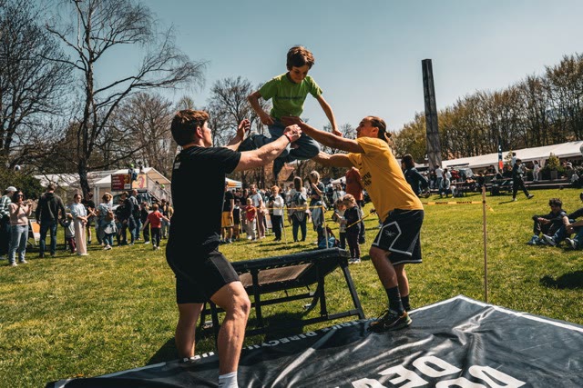 Child springing high above a mini trampoline, supported by two coaches, at a family event