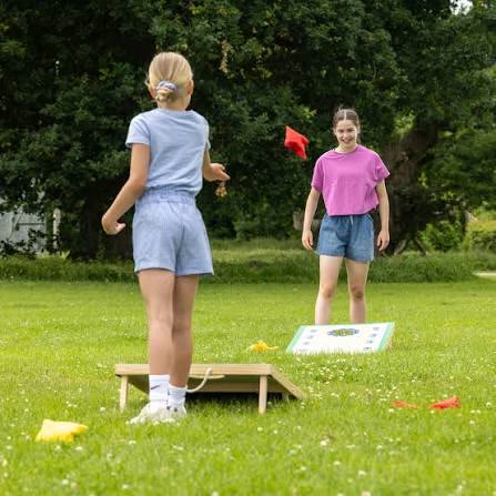Two girls playing cornhole on a lawn, one tossing a red bag towards the board