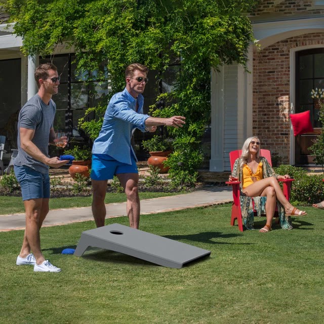 Friends playing cornhole in a garden while others watch