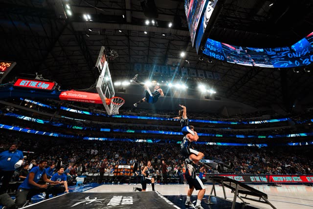 Athlete in a tower dunk over teammates and a trampoline ramp in a large basketball arena