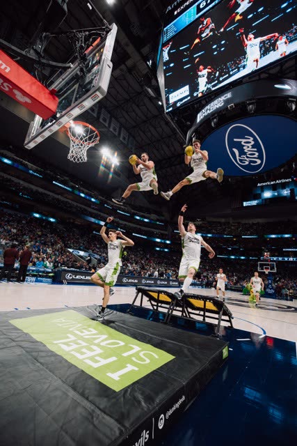 Two athletes in white kits dunking side by side high above the court in an NBA arena