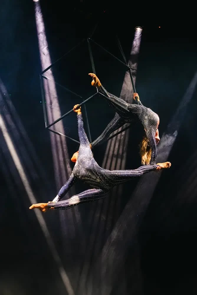 Two aerialists in sparkling costumes performing on a suspended geometric aerial cube in spotlight beams