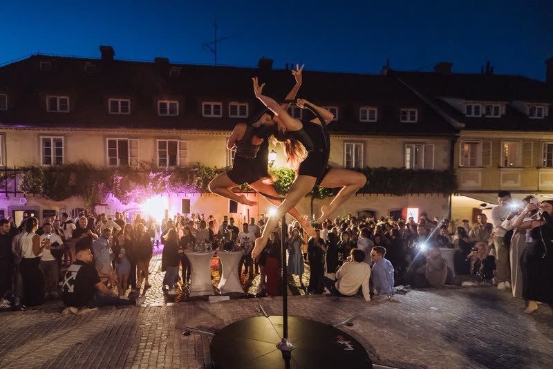Two aerialists on a freestanding aerial hoop at a night-time outdoor event in a town square with an audience