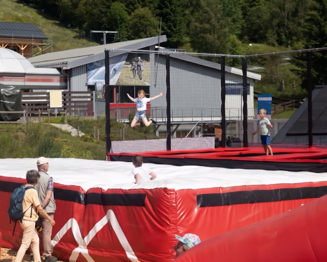 Child jumping off the edge of a large red and white airbag next to a trampoline court at a family event