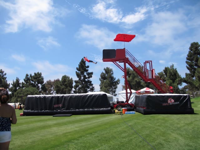 Participant leaping from a red tower towards two black airbags on a lawn at an outdoor event