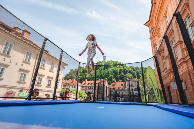 Child jumping high on a blue trampoline inside a safety net with Ljubljana old town and castle hill behind