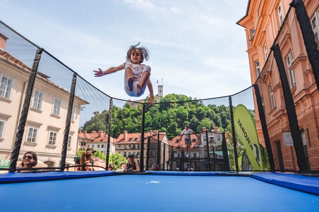 Child flying through the air above a blue trampoline surrounded by a safety net at a city event