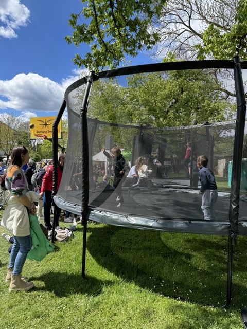 Children bouncing on a round trampoline with a safety net in a park next to a Dunking Devils hoop