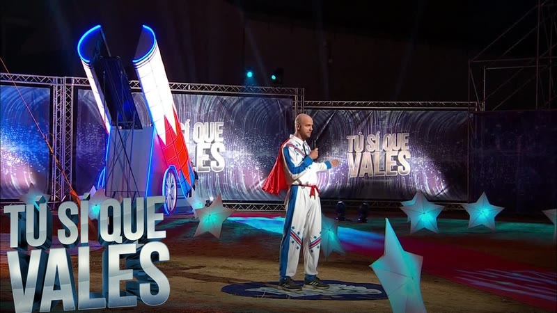 Performer in a white and blue stunt suit with a red cape standing in front of the illuminated human cannon on a night stage
