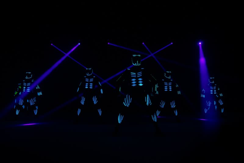 Five LED dancers in blue glowing costumes standing in a row on a dark stage under crossing purple light beams