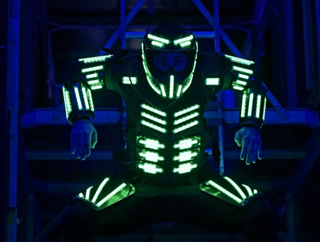 Close-up of an LED dancer in a helmet and green glowing costume against a blue metal structure