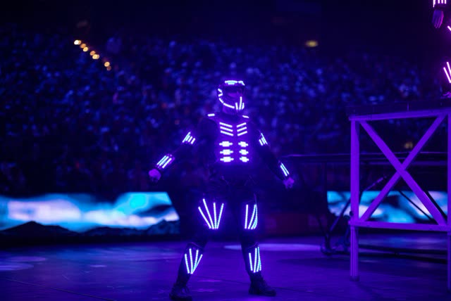 LED dancer in a violet glowing costume on a large arena stage in front of a packed audience
