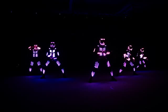 Five LED dancers in pink and purple glowing costumes performing a choreography on a dark stage