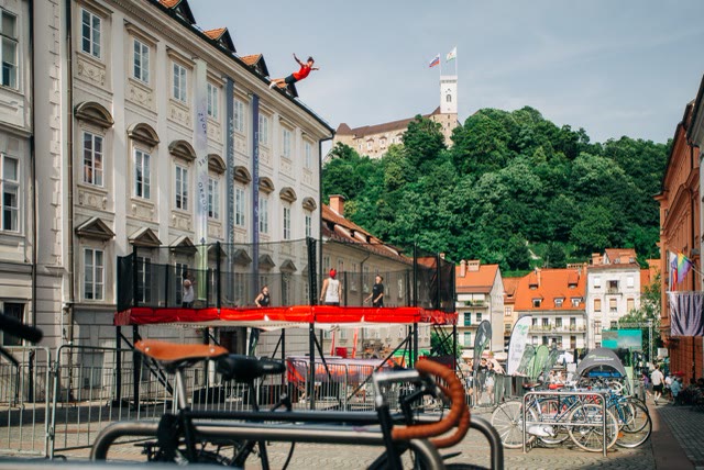 Raised trampoline with red padding and safety nets in a Ljubljana street with an acrobat flying above it and the castle hill behind