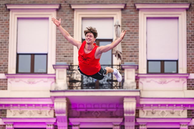 Smiling acrobat in a red vest in mid-air in front of a pink-lit building