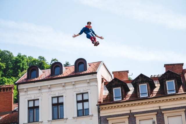 Performer in a superhero costume flying above old town roofs against a blue sky