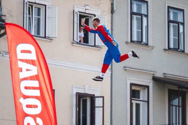 Performer in a red and blue costume leaping past a banner with a spectator waving from a window