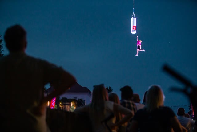 Performer hanging from a cable below a punching bag at dusk while a crowd watches