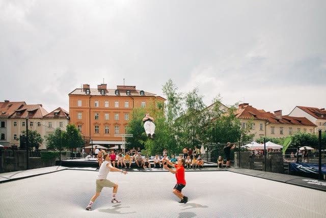 Wide view of the giant trampoline on a historic square with a jumper in mid-air and spectators sitting at the edge