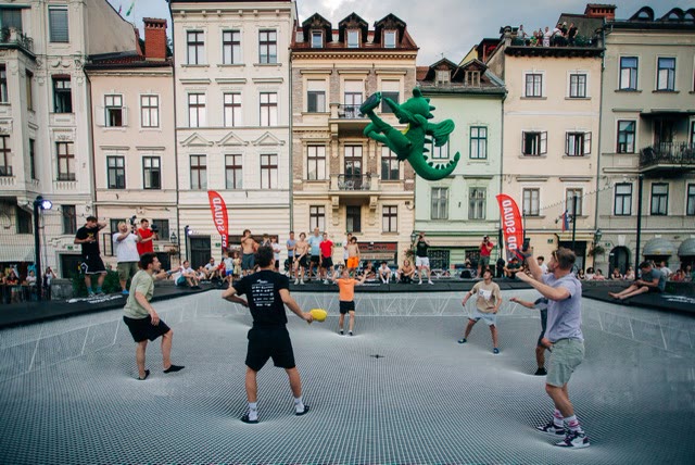 People playing a ball game on the giant trampoline in a city square with spectators and a green inflatable dragon above