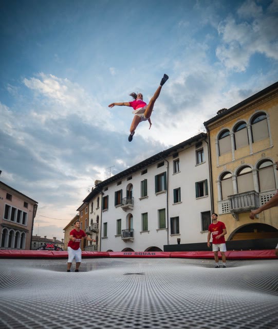 Girl in a split jump high above the trampoline in a town at sunset with two coaches below