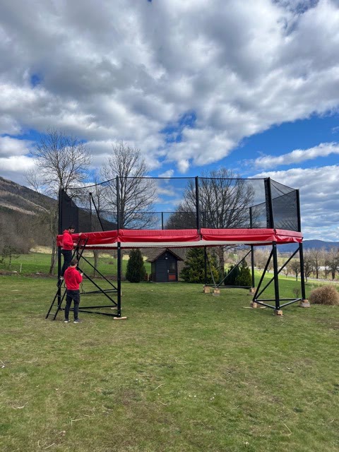 Raised megatramp with red padding and safety nets being set up on a lawn, with a coach on the ladder