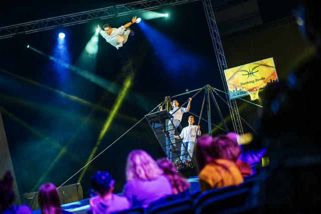 Acrobat flying high above the audience from a russian swing in blue stage light with a Dunking Devils banner behind