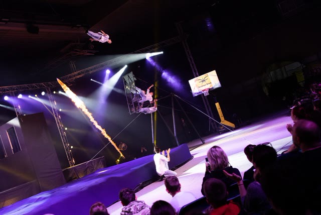 Acrobat leaping off the russian swing above an airbag stage with a flame effect and an audience in the foreground