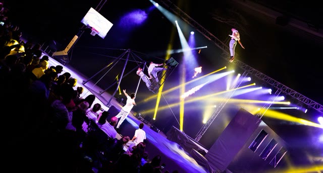 Performers launching from the russian swing in stage lights with flame effects and spectators on the left