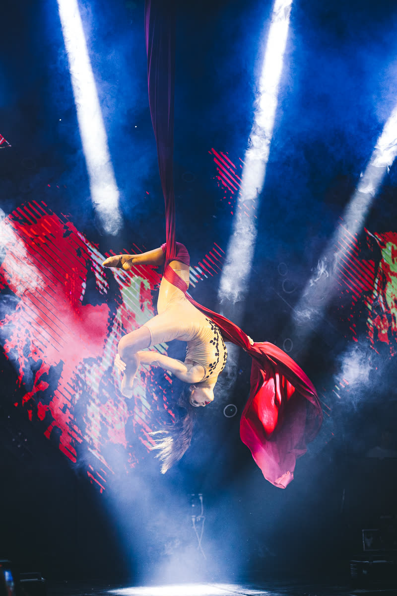 Aerialist upside down on red aerial silks in blue light beams and stage smoke