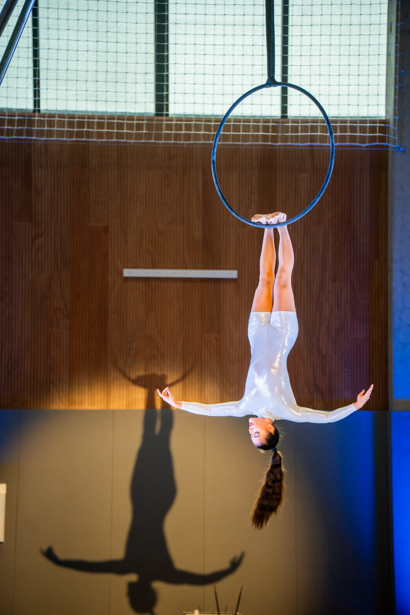 Aerialist hanging upside down from an aerial hoop in a sports hall, casting a long shadow