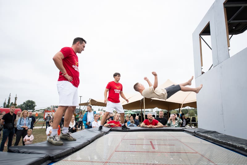 Boy doing a back flip above a trampoline while two coaches in red shirts spot him beside the trampoline wall