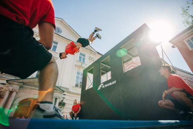 Boy flipping in mid-air above a trampoline in front of a dark trampoline wall in a historic town square