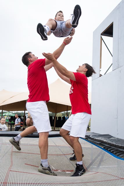 Two coaches in red shirts tossing a boy high above a trampoline beside the trampoline wall