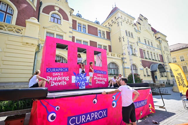 Children sitting in the openings of the pink trampoline wall on a town square with spectators around
