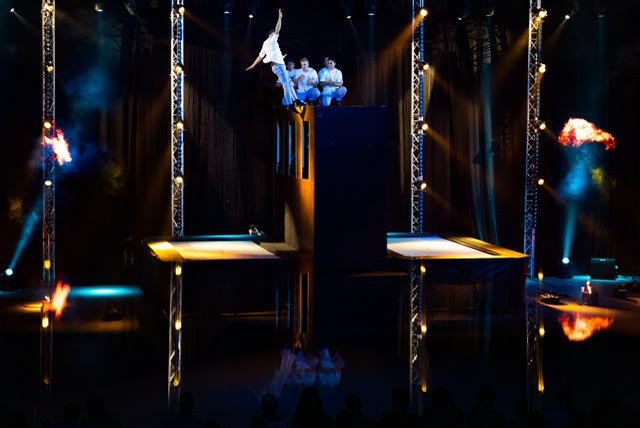 Acrobat leaping from the trampoline wall while three performers sit on top, with reflections on the stage floor