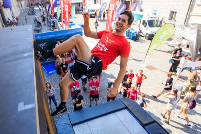 Smiling Dunking Devils acrobat in a red shirt hanging on the edge of the trampoline wall above a city street crowd