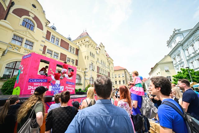Acrobats sitting in the window openings of a pink trampoline wall on a town square with spectators
