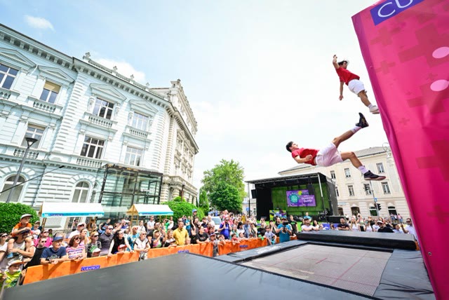 Two acrobats in mid-air above the trampoline beside a pink wall with a large crowd around the stage