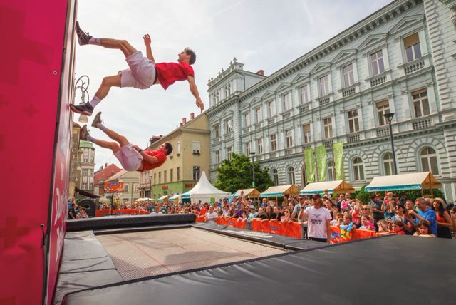 Two acrobats flipping side by side next to the pink trampoline wall at a city festival